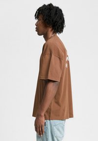 Brown cotton t-shirt with short sleeves, relaxed fit, and a crew neckline; logo printed on the back in white. Worn with light blue jeans.