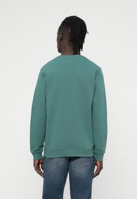 Πράσινο φούτερ με στρογγυλή λαιμόκοψη, μακριά μανίκια και ριμπ τελειώματα. Φορεμένο με μπλε τζιν, δείχνοντας μια ομοιόμορφη, απαλή υφή.