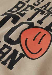 Bomuldsstof med fed sort tekst og et orange smilende ansigt grafisk motiv på en beige baggrund, der formidler et legende design.