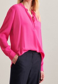 Blusa fucsia a maniche lunghe con scollatura a V e vestibilità rilassata. Tessuto liscio, dettaglio con bottoni sul collo, abbinata a pantaloni scuri.