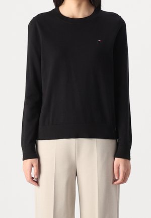 Frau trägt einen eng anliegenden schwarzen Pullover mit langen Ärmeln und lockere beige Hose, steht vor einem schlichten hellen Hintergrund.