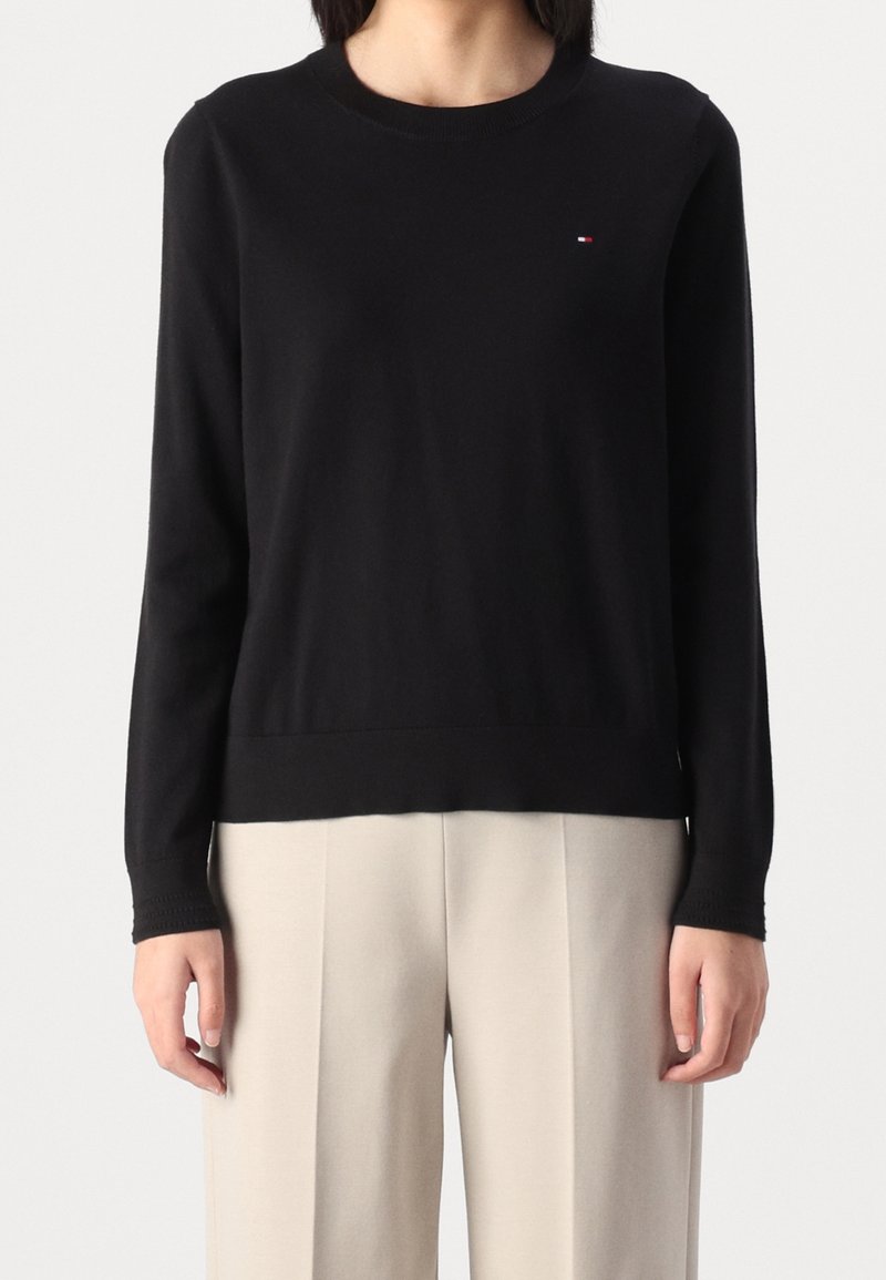 Femme portant un pull noir ajusté à manches longues et un pantalon beige ample, debout devant un fond clair uni.