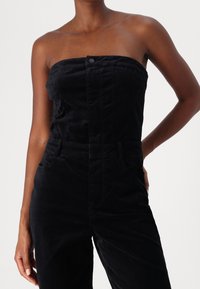 Svart ärmlös jumpsuit i mjukt, texturerat tyg. Har en knappstängning, fickor och en figursydd silhuett.