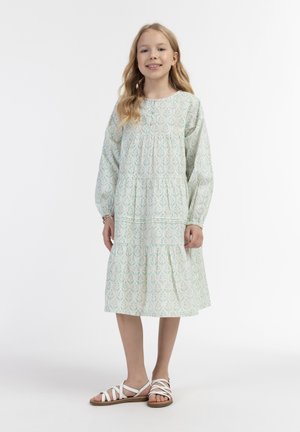 Lys pastellgrøn kjole med blomster mønster, lagdelt design, lange ærmer og rund halsudskæring, parret med hvide, stroppede sandaler.