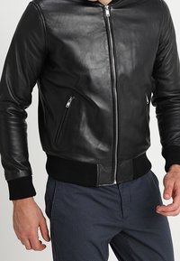 Svart läder bomberjacka med dragkedja fram, ribbade ärmslut, två fickor med dragkedja och en jämn textur. Har en figurnära design.