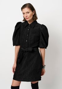 Zwarte jurk met opbollende mouwen, knoopsluiting en twee voorzakken. Gestructureerde stof en een strikceintuur accentueren de taille.