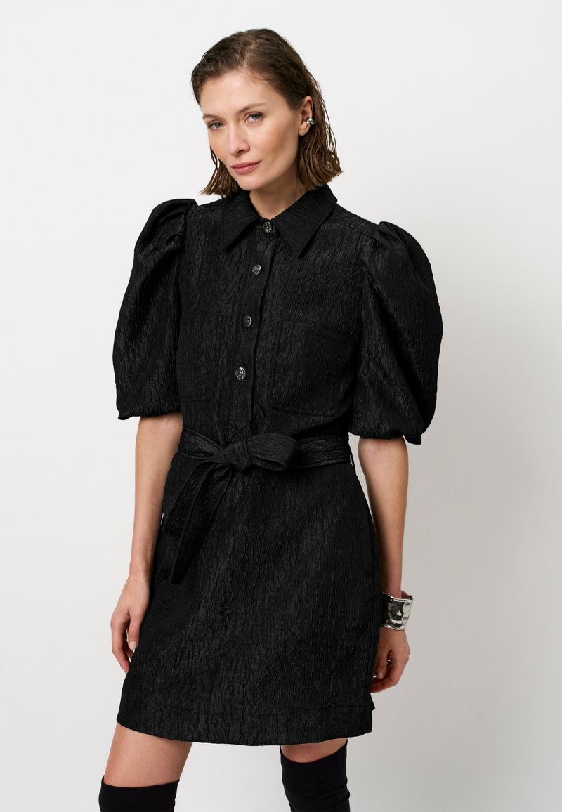 Zwarte jurk met opbollende mouwen, knoopsluiting en twee voorzakken. Gestructureerde stof en een strikceintuur accentueren de taille.