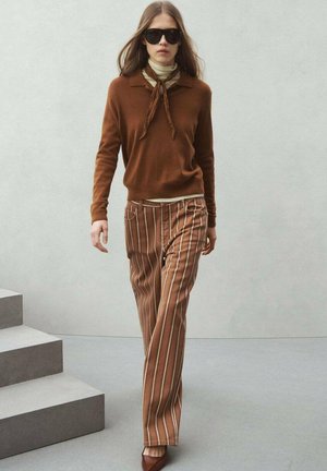 Mujer caminando con suéter marrón, cuello alto crema, pantalones marrones a rayas, zapatos puntiagudos y gafas de sol negras sobre un fondo liso.