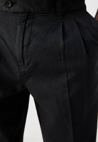 Black trousers featuring a textured fabric, pleated front, and front pockets. The waistband includes a button closure for secure fit.