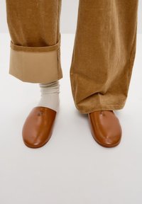 Scarpe slip-on in pelle marrone con punta rotonda, dotate di un piccolo logo metallico. Indossate con calzini chiari e pantaloni di velluto a coste con risvolto.