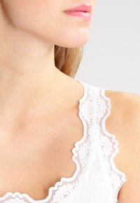 Top bianco senza maniche con bordo in pizzo scallop lungo il colletto, realizzato in un tessuto a costine, con la pelle liscia visibile sotto.