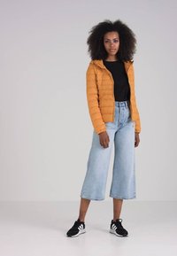 Oransje puffjakke med hette, svart skjorte under, høyhattet lys blå vide jeans og svarte joggesko med hvite detaljer.