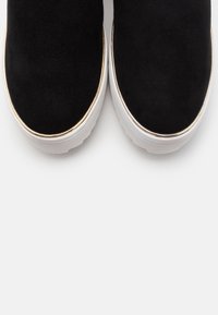 Czarne zamszowe buty wsuwane z zaokrąglonym noskiem, z białą gumową podeszwą i złotym akcentem wokół górnej krawędzi.