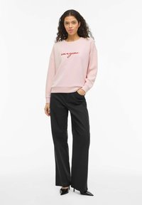 Lyserosa genser med "imagine" brodert i rødt, kombinert med svarte bukser med vide ben og svarte spisse hæler. Jevn stofftekstur.