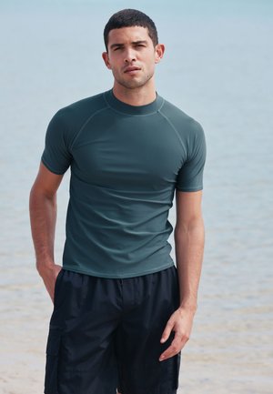 Jauns vīrietis, kurš valkā tumši zaļu rash gardu ar īsām piedurknēm un melnas peldbikses, stāv pludmalē ar ūdeni fonā.