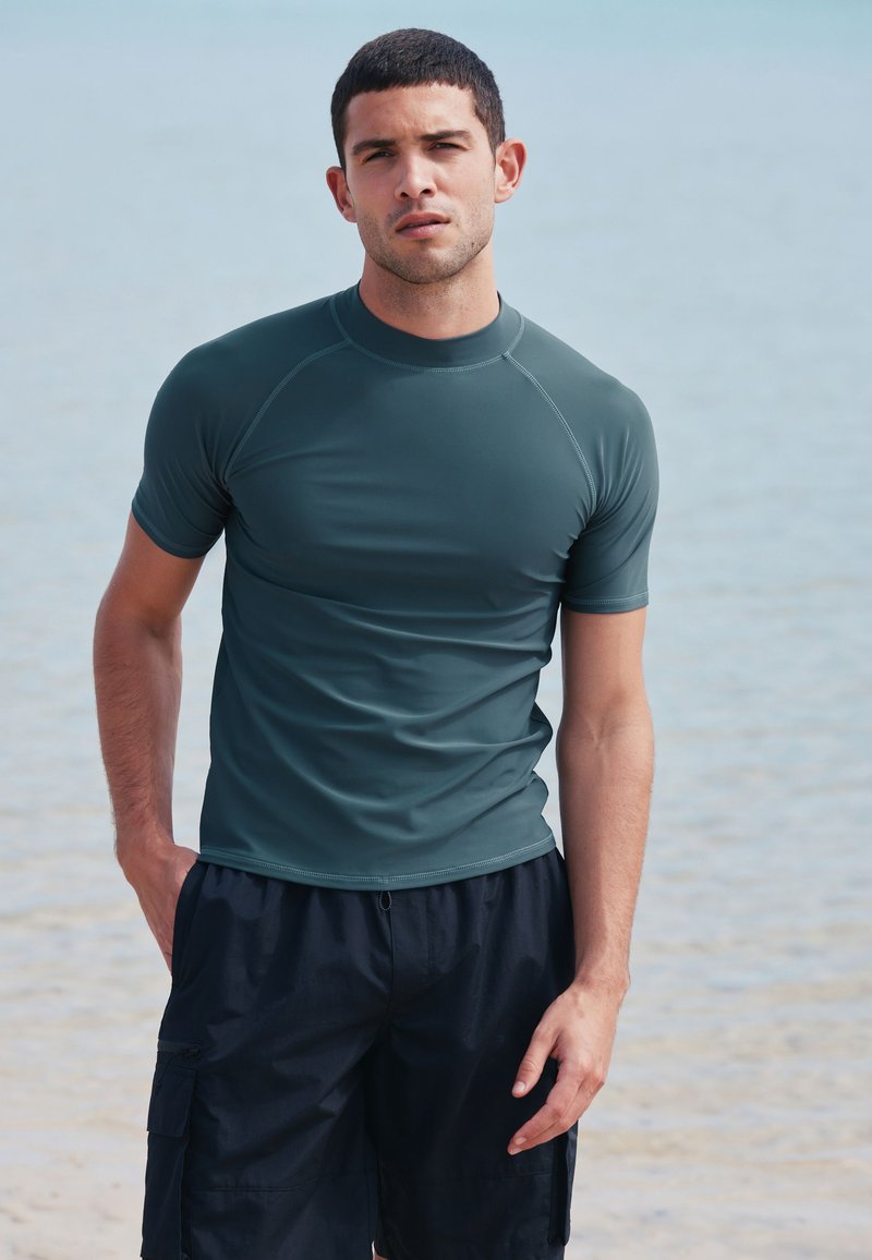 Jeune homme portant un rash guard à manches courtes vert foncé et un short de bain noir, debout sur une plage avec de l'eau en arrière-plan.