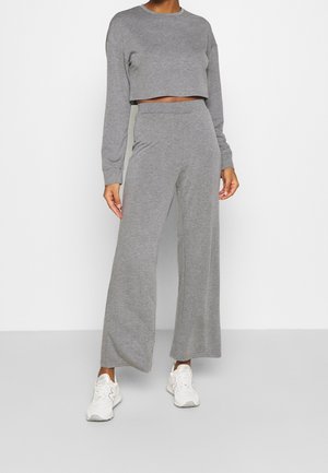 Personne portant un crop top gris à manches longues et un pantalon large assorti, avec des baskets blanches, debout devant un fond blanc uni.