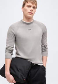 Jeune homme portant un pull BOSS gris clair et un pantalon noir, tenant une pochette noire de la marque Hugo sur un fond clair uni.