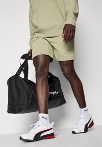 Sportieve set met een lichtolijfgroen sweatshirt, bijpassende shorts, witte en zwarte PUMA sneakers en een zwarte sporttas met witte opdruk.