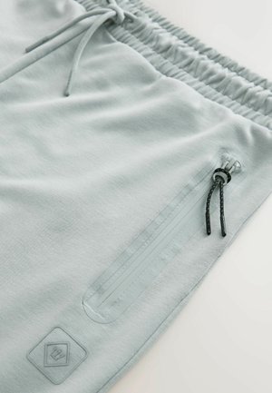 Un pantalon de jogging gris clair avec un tissu texturé, une taille élastique, une poche zippée avec des accents noirs et un cordon de serrage ajustable.