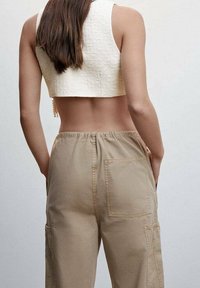 Femme aux longs cheveux bruns portant un crop top blanc sans manches et un pantalon taille haute beige avec des poches, de dos par rapport à l'appareil photo.