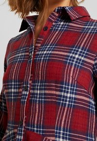 Camicia a quadri rossa e blu con bottoni, dotata di colletto e bottoni neri, realizzata in un morbido tessuto di flanella a trama. Vista ravvicinata del motivo.