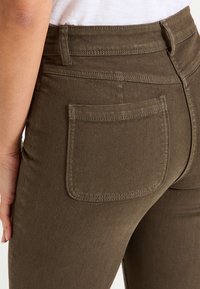 Pantalon en denim vert foncé au design ajusté, taille haute, et une poche arrière avec des surpiqûres contrastées. Texture lisse et lignes épurées.