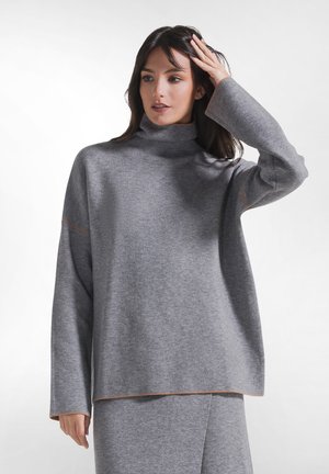 Kvinde iført en løstsiddende grå rullekravesweater og matchende nederdel, med den ene hånd, der rører ved hendes hår, mod en ensfarvet hvid baggrund.