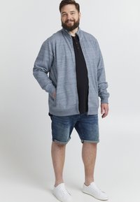Pelēks zip-up džemperis ar teksturētu audumu, melna polo krekls zem tā, džinsus šorti un baltas kedas. Modelis ir ar kabatām džemperī.