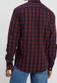 Mies, który ma na sobie koszulę w czerwono-czarną kratę z długim rękawem, zestawioną z niebieskimi dżinsami, widoczny z tyłu na jednolitym tle.
