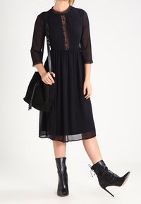 Schwarzes Kleid mit Polka-Dots, drei Viertelärmeln und Spitze am Ausschnitt, kombiniert mit schwarzen Stiefeletten und einer schwarzen Tasche.