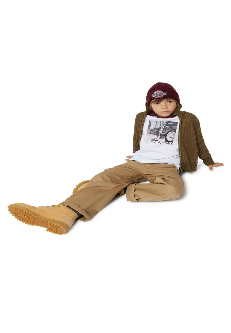 Dziecko ubrane w brązowy żebrowany kardigan, biały t-shirt z grafiką, beżowe spodnie i brązowe buty z wytrzymałą podeszwą. Na głowie ma burgundową czapkę.