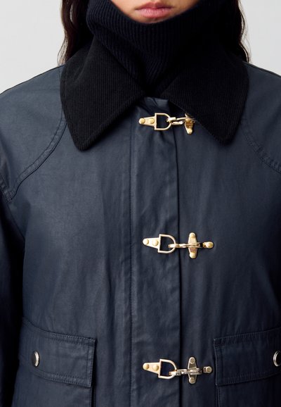 Veste imperméable bleu marine avec un col en ribbed noir, des fermetures à glissière dorées et des poches latérales. Tissu texturé avec une finition lisse.