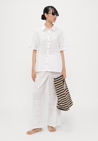 Conjunto de tela de ojo de perdiz blanca con camisa de manga corta, pantalones holgados y botones. La modelo sostiene un bolso a rayas. Sin zapatos. Textura ligera y transpirable.