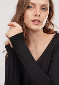 Schwarzer Strickpullover mit V-Ausschnitt und gerippten Bündchen. Weiche Textur mit enger Passform. Das Modell zeigt die Handposition in der Nähe des Ausschnitts.