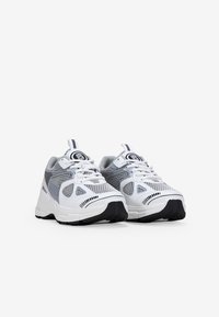 Zapatillas deportivas blancas y grises con una parte superior de malla, suela negra gruesa, diseño de cordones y acento de logo en la lengua.