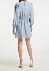 Frau in einem hellblauen Langarmkleid mit gerüschter Taille und Bündchen, stehend, von hinten gezeigt, trägt weiße High-Heels.
