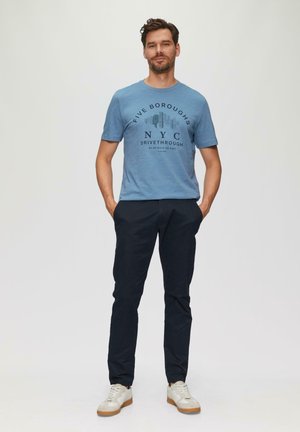Lichtblauwe t-shirt met korte mouwen met de graphic "FIVE BOROUGHS NYC DRIVETHROUGH", gecombineerd met donkerblauwe broek en witte sneakers.