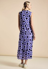 Vestido maxi en púrpura con un patrón geométrico, diseño sin mangas, cintura fruncida y falda en capas; combinado con zapatillas blancas.