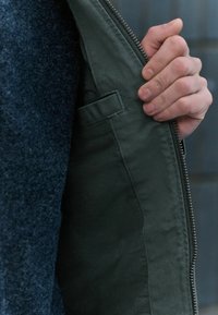 Veste verte avec fermeture éclair et intérieur texturé, présentée partiellement ouverte. Dispose d'une petite poche à l'intérieur.