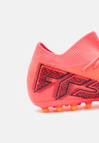 Botín de fútbol de color rosa brillante con un upper texturizado, presentando acentos gráficos en negro y tacos naranjas en la suela para tracción.