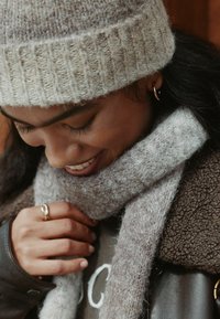 Light gray wool knit beanie with ribbed texture; matching light gray wool scarf; hand with gold ring resting on dark brown shearling jacket.