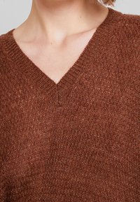 Pull en bronze tricoté avec un col en V, un motif texturé et des finitions côtelées. Le matériau présente un léger éclat et une finition douce.