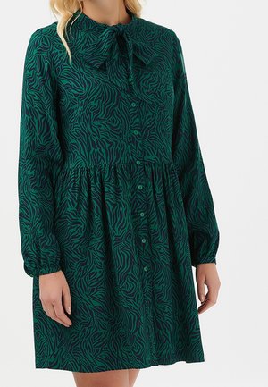 Mujer que lleva un vestido verde oscuro con rayas negras de cebra, mangas largas abullonadas, botones en la parte delantera y un cuello con lazo.