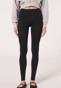 Schwarze Leggings mit hohem Bund, aus einem glatten, dehnbaren Stoff gefertigt. Sie haben ein figurbetontes Design und einen knöchellangen Schnitt, kombiniert mit schwarzen Ballerinas.