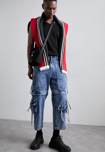 Cardigan roșu și gri cu dungi, tricou negru, blugi cargo albaștri cu șnururi și rupturi, și adidași negri.