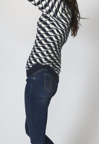 Svetr pletený s námořnickými a bílými vlnovkovými vzory s texturovaným povrchem, kombinovaný s tmavě modrými pružnými džíny s dekorativními zadními kapsami.