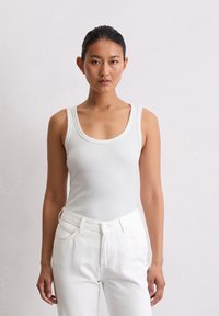 Weißes, geripptes Tanktop mit einem Rundhalsausschnitt, kombiniert mit hochgeschnittenen weißen Jeans. Der Stoff wirkt weich und strukturiert.