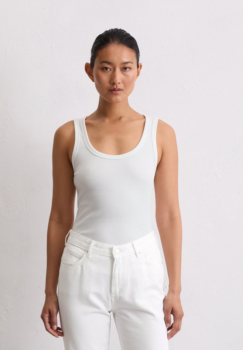 Weißes, geripptes Tanktop mit einem Rundhalsausschnitt, kombiniert mit hochgeschnittenen weißen Jeans. Der Stoff wirkt weich und strukturiert.