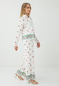 Ein Model läuft in einem weißen, floral bestickten Outfit, das mit bunten Blumen und einem gemusterten Saum verziert ist, kombiniert mit weißen, schulterfreien Absätzen.